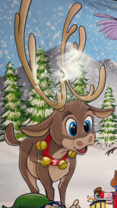 Close-up artwork of a cute, wide-eyed reindeer with large antlers and a red collar with gold bells, set against a snowy mountain and pine tree backdrop.