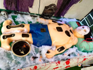 A Halloween-themed operation game (similar to Operation or Sawbones) featuring a large plastic body wearing blue shorts, lying on fake spiderwebs and a blood-spatter cloth. The body has holes containing game pieces, is surrounded by toy spiders, and includes a creepy mask, a tombstone prop, and a digital score counter.