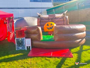Full view of the Mechanical Rodeo Pumpkin ride set up on grass, surrounded by an inflatable, wood-look safety barrier. A separate red tent and a control box with Halloween signage are visible on the left.