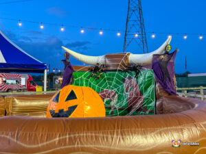 Angled view of the Rodeo Pumpkin setup. The inflatable ring contains the ride, and the background shows surrounding event stalls, including a food bar and a blue and white tent.