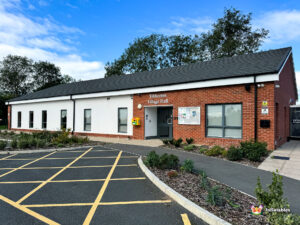 Wider exterior view of Tibberton Village Hall, showing the entrance and surrounding landscaped beds and the car park markings in the foreground.