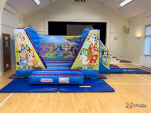 Wide view of the Bluey Bounce and Slide inflatable, showing the full structure set up on the wooden floor of a hall, with safety mats placed around the entrance and exit.