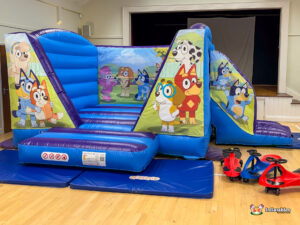 Angled view of the entire Bluey Bounce and Slide inflatable, clearly showing the entrance and the blue and purple slide exit on the right, with three red and black ride-on toys next to the exit mat.