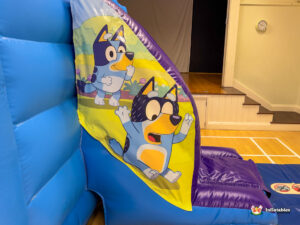 Close-up of the art near the slide entrance, featuring two dynamic illustrations of the character Bluey with an exaggerated, happy expression, right before the blue and purple slide ramp.