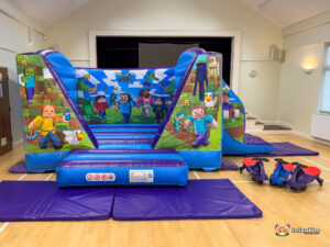 Wide frontal view of the complete Minecraft Bounce and Slide inflatable set up in a hall. The entrance pad and surrounding purple safety mats are visible, along with three small ride-on toys on the right.