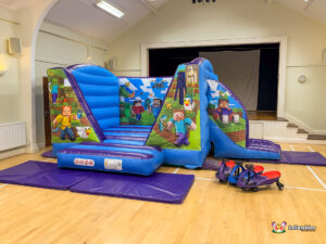 Wide view of the complete Minecraft Bounce and Slide inflatable set up in a hall. The blue and purple structure, purple safety mats, and three small ride-on toys are visible on the wooden floor.