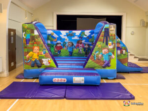 Frontal view of the complete Minecraft Bounce and Slide inflatable set up on purple safety mats, showing the character art on the columns and the back wall, framed by the hall's entrance.