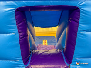 Interior close-up looking through a purple-lined exit hole of the blue inflatable, showing the safety mat below and a small warning label on the floor.