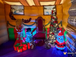 Interior scene of the grotto Photo Pod showing a red throne, a large reindeer prop, a lit Christmas tree, and light-up Santa and snowman figures, all against the inflatable walls decorated with winter paintings.