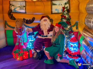 Santa Claus seated inside the grotto, posing next to a smiling female Elf who is kneeling on the floor. They are surrounded by light-up gifts, a large reindeer prop, and other festive decorations.