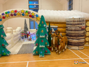 Wide exterior view of the full attraction, showing the inflatable log cabin, the cylindrical Santa's Photo Cabin, and the inflatable archway leading into a separate snowy walk-through scene.