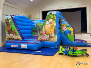 Wide angled view of the inflatable set up in a community hall, showing the slide exit on the right and three colorful ride-on cars in the foreground.