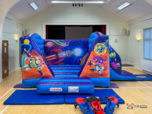 A frontal view of a blue and purple space-themed inflatable bounce house and slide set up indoors, featuring Buzz Lightyear and various cartoon alien characters.
