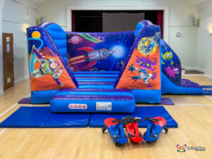 A wide shot of the space-themed inflatable unit in a hall, including three colorful ride-on toys in the foreground and a visible slide exit on the right.
