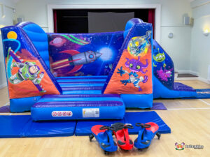 A full frontal view of the inflatable unit, showcasing the complete set of space murals and characters, set up with safety mats and wiggle cars on a wooden floor.