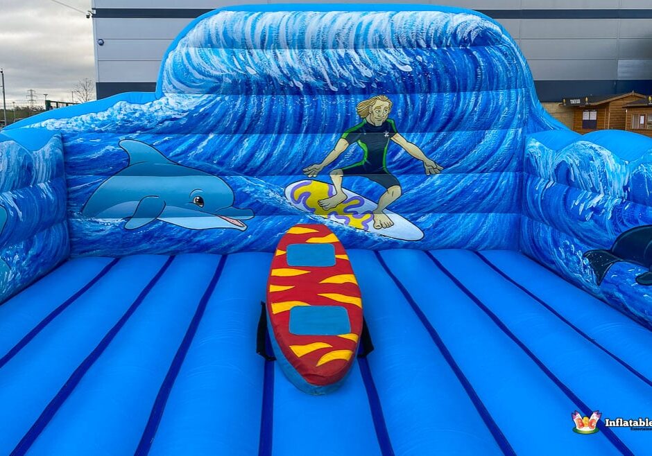 A colourful inflatable surf-themed simulator with a surfer, dolphins, and wave patterns, featuring a red surfboard in the centre.
